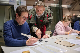 Besucher probieren in den Zwickauer Priesterhäusern an einem Tisch unter Anleitung eines historisch gekleideten Mitarbeiters das Schreiben mit Feder und Tinte aus.
