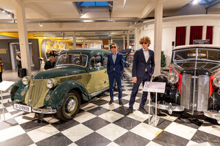 Zwei Männer in Anzügen und Sonnebrillen stehen zwischen zwei historischen Automobilen im August Horch Museum Zwickau.